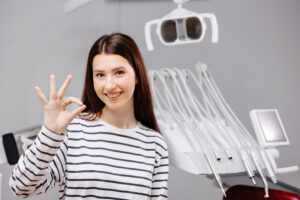 Smiling woman in a striped shirt shows an 'OK' gesture in a dental office, surrounded by dental equipment, conveying a positive and welcoming atmosphere.