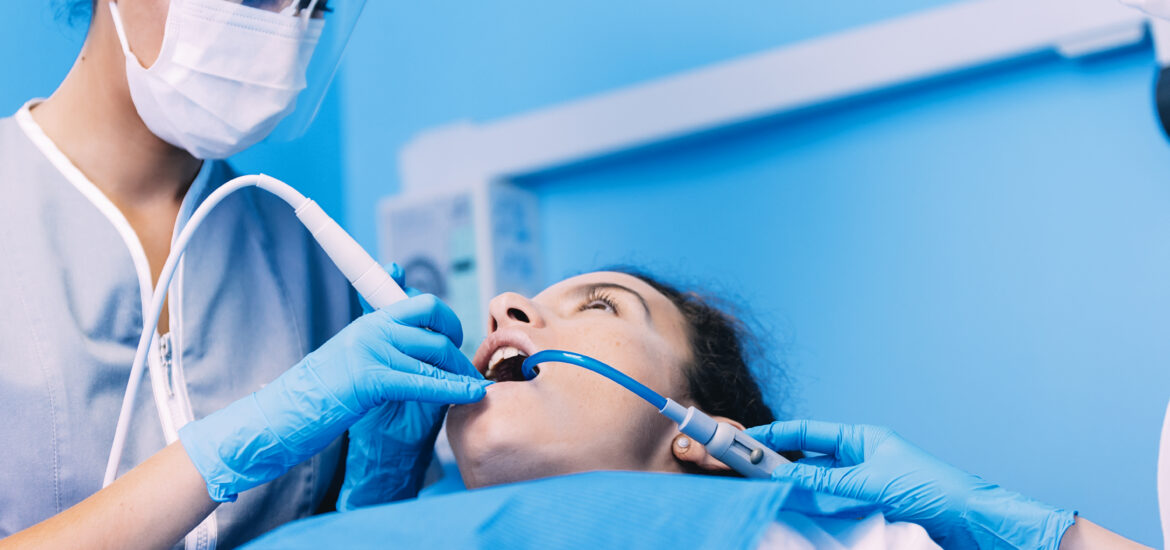 Dentist in protective gear and mask uses dental tools on a patient in a blue dental clinic. The atmosphere is professional and clinical.