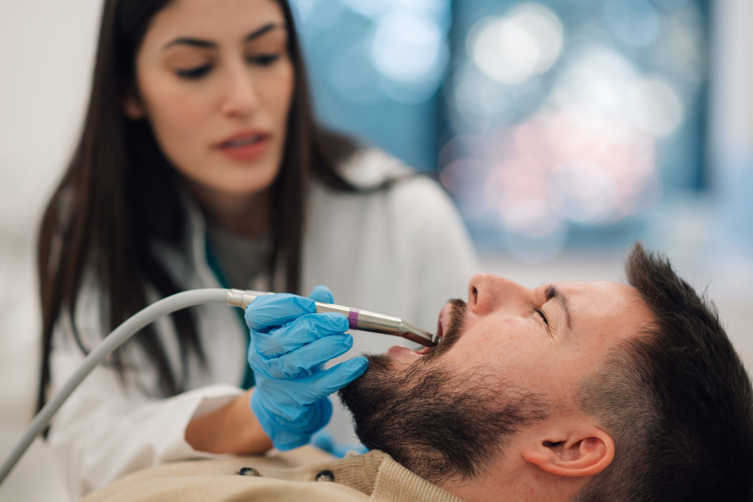 A dentist wearing blue gloves examines a patient's mouth with dental tools. The patient is lying back in a chair, and the setting is a bright, modern clinic.